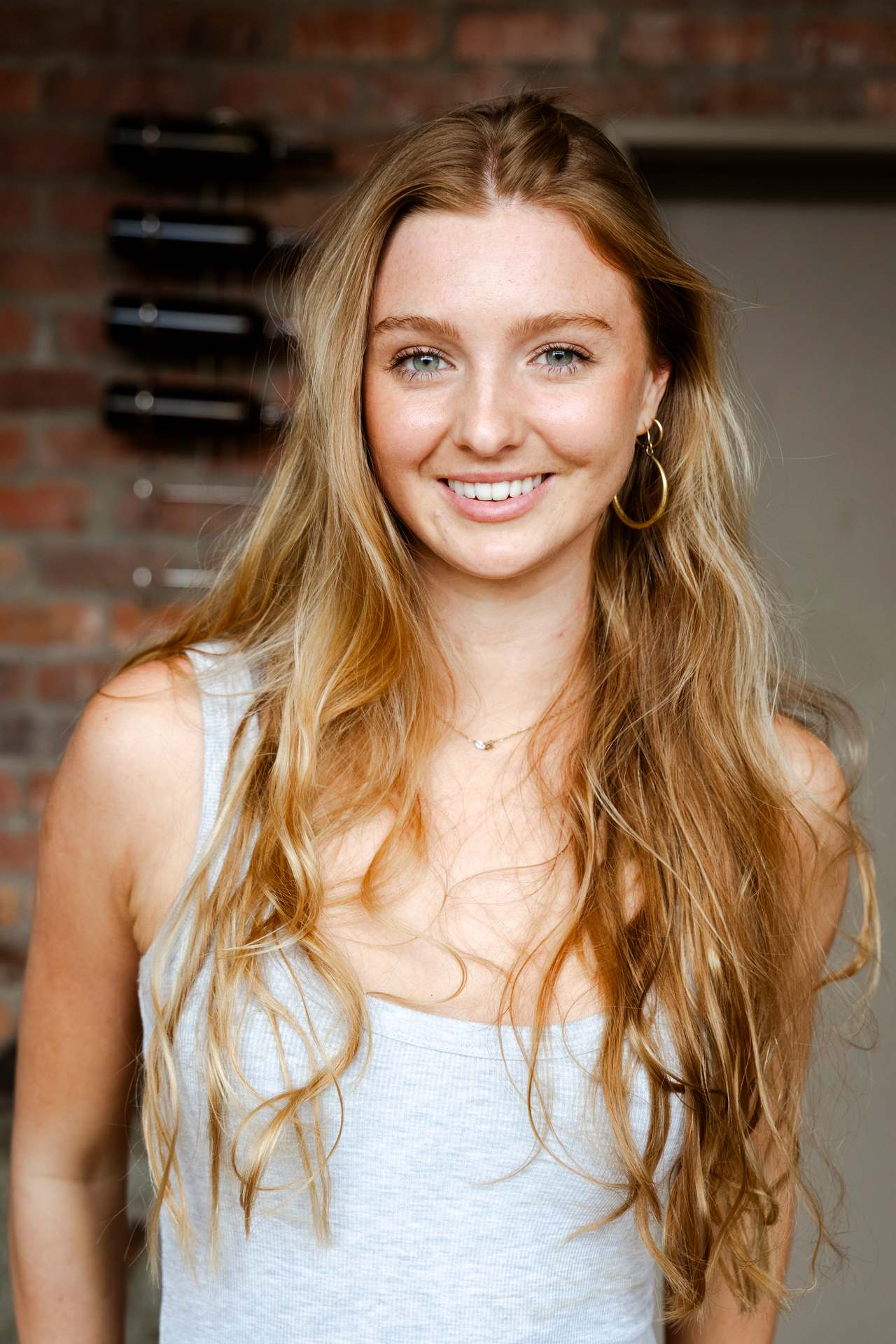 Digitals headshot: Georgia Bauriedl smiling, hair down, grey vest top, brick wall behind.