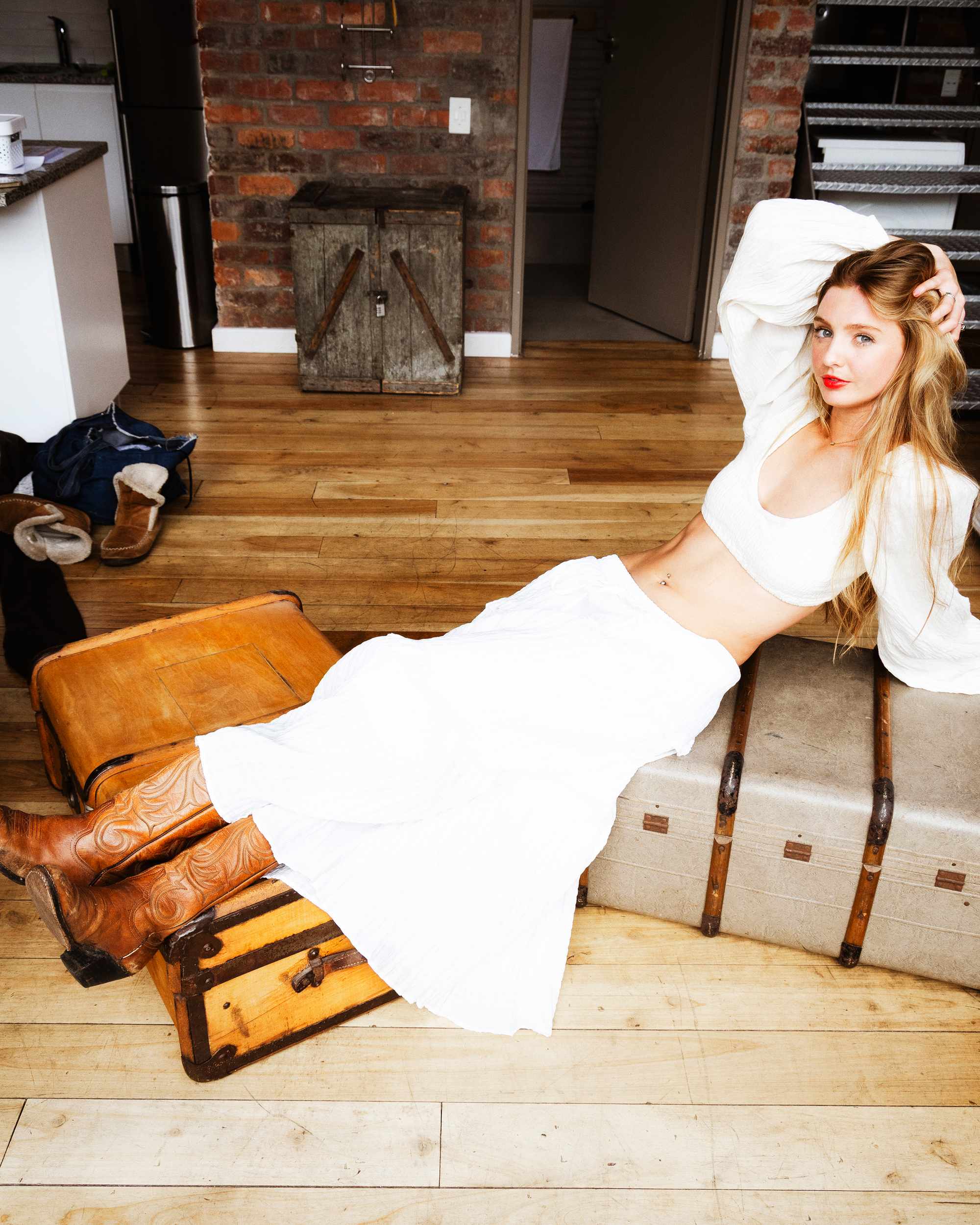 Georgia Bauriedl reclining across vintage travel trunks in a white skirt and top, cowboy boots beside her.